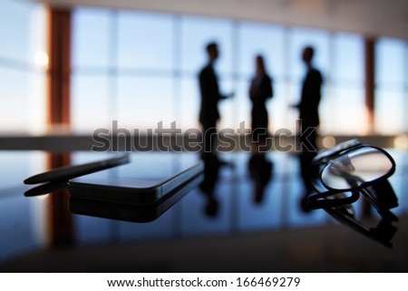 Close-up of eyeglasses, cellular phone and pen at workplace on background of office workers interacting Royalty-Free Stock Photo #166469279