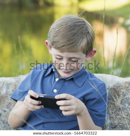 A young boy outdoors sitting leaning against a rock, using a handheld electronic game.