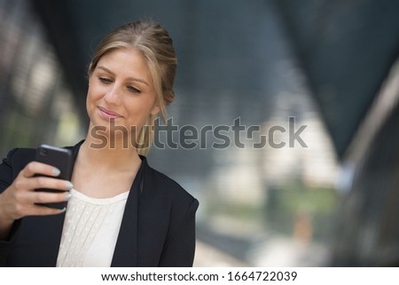 A young blonde businesswoman on a New York city street. Wearing a black jacket. Using a smart phone.