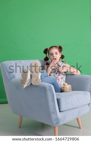 Cool naughty girl of seven years old with a funny hairstyle sitting in a big blue armchair and eating crunchy potato chips on a bright green background