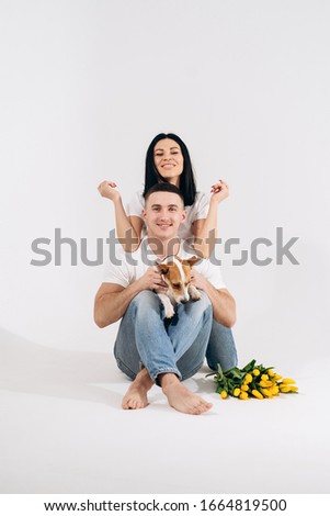 Close up portrait young couple sit and hugging, holding yellow flowers and dog in studio on white background. couple embracing with dreamy amorous expression. Lovely family. Celebrating woman's day.
