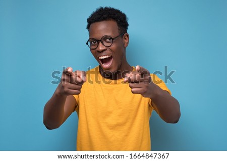 Handsome young african man in yellow clothes and glasses pointing his finger at you standing on blue wall. Studio shot.