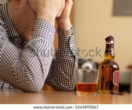 drunk man sitting at the table holding his head. on the table is a glass with alcohol and an open bottle. problem with alcohol