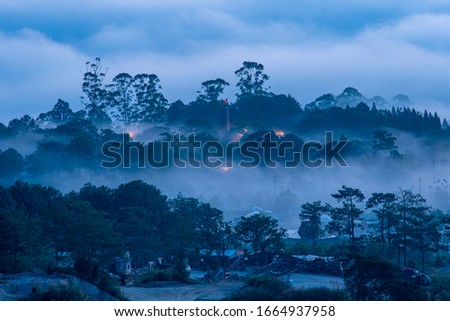 Mountains in fog at beautiful morning in autumn in Dalat city, Vietnam. Landscape with Langbiang mountain valley, low clouds, forest, colorful sky , city illumination at dusk.