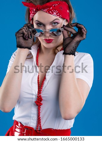 An attractive girl with a red bandana on her head attentively looks with lowered glasses. Girl in pin-up style isolated on blue background