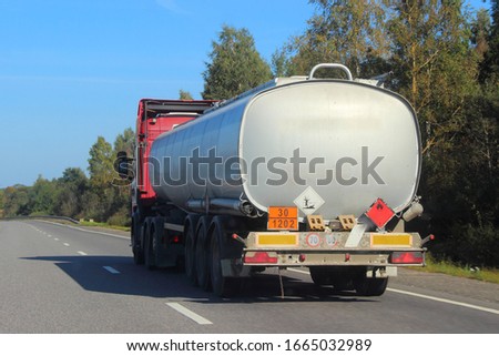 Oil transportation, silver gray fuel tank truck with ADR sign 30 1202 on suburban asphalted road at Sunny summer day on blue sky and green trees background, flammable liquid transportation logistics