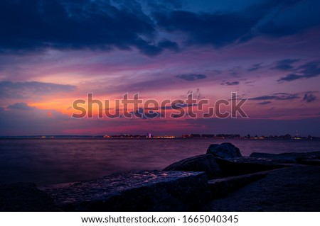 Watching the sunset on Breezy Point Beach on the end of one summer warm day. Dramatic cloudy dark blue sky and orange purple touch of the sun in the background. Big rocks in the foreground.