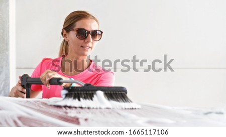Young woman wearing pink t shirt and sunglasses cleaning her car in self serve carwash, spreading foam with brush over roof, banner with space for text right side