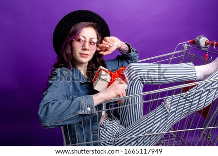 Stylish girl in jeans with pink glasses sitting in a grocery cart on a purple background