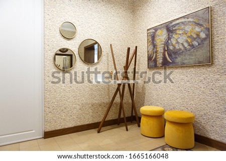 The interior of the hallway in a suburban house. In the hallway there are elements: yellow ottomans, pigeon-shaped hangers, figurines, a picture, a chandelier. Entrance to the main room.