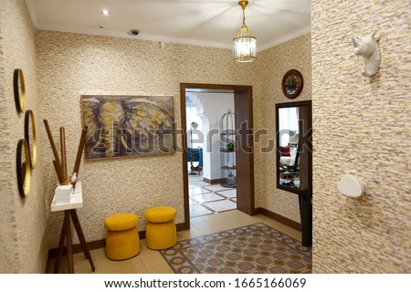 The interior of the hallway in a suburban house. In the hallway there are elements: yellow ottomans, pigeon-shaped hangers, figurines, a picture, a chandelier. Entrance to the main room.