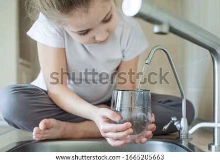 Little girl open a water tap with her hand holding a transparent glass. Kitchen faucet. Filling cup beverage. Pouring fresh drink. Hydration. Healthcare. Healthy lifestyle. World Water Day Royalty-Free Stock Photo #1665205663