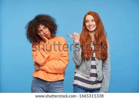 Indoor shot of cheerful lovely redhead female showing happily with thumb on her upset curly dark skinned brunette girlfriend whileposing over blue background