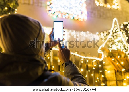 Rear view of man holding blank screen mobile phone against house decorated with christmas lights.