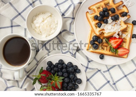Beautiful breakfast: Viennese Belgian waffles decorated with berries, a plate with strawberries and blueberries, a bowl with coconut flakes, a mug of black coffee and cutlery on a checkered napkin