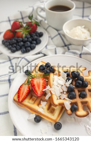 Beautiful breakfast: Viennese Belgian waffles decorated with berries, a plate with strawberries and blueberries, a bowl with coconut flakes, a mug of black coffee and cutlery on a checkered napkin