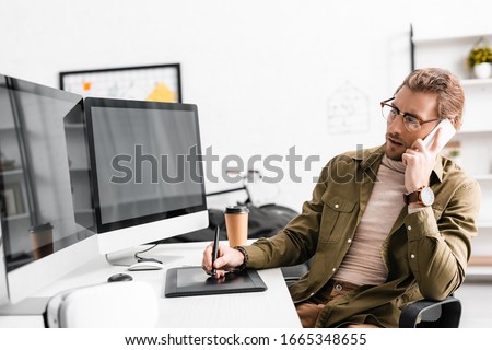 3d artist talking on smartphone and using graphics tablet and computers at table in office