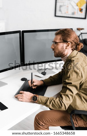 Side view of handsome digital designer working with graphics tablet and computers with blank screen on table