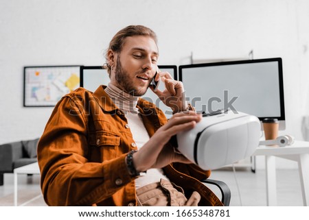 Selective focus of handsome digital designer talking on smartphone and holding virtual reality headset in office