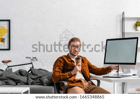 Pensive digital designer with paper cup holding stylus near graphics tablet and computer on table