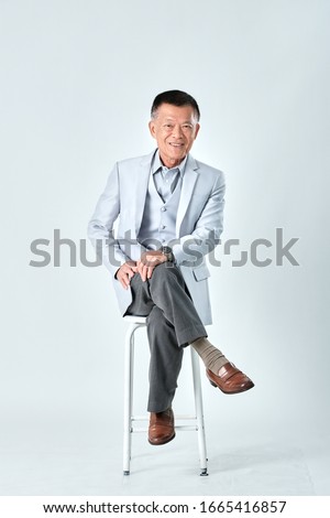 Full body of an old asian man wear light grey suit sitting on a grey chair and cross one's legs in studio white background. Isolated picture of happiness asian old man.