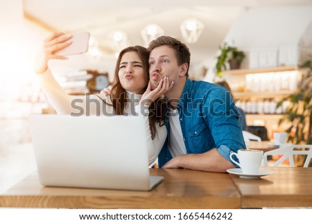 young couple beautiful woman and handsome man grimacing shooting selfie on smarthone while drinking coffee during lunch time break at cafe, internet technology concept