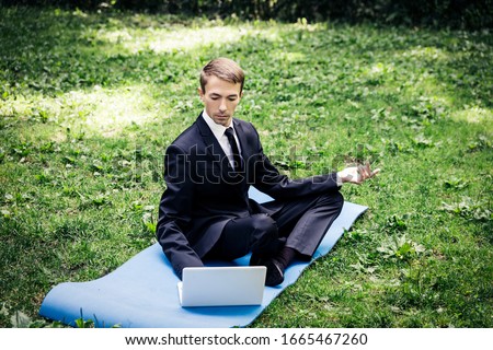 Man in a suit with tie. Business man relaxing in a park in the lotus position, he can't relax in any way, typing text on a computer with one hand. Looks  into the monitor.