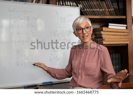 Portrait of senior professor explaining  math formulas. Mature female teacher standing in classroom and talking to her class. Lecturer explaining math formulas written on whiteboard in library. Royalty-Free Stock Photo #1665468775