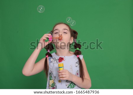 Funny girl of eight years old with a funny hairstyle in a blue blouse with flowers fiddles a bottle in her hands and blows soap bubbles children's entertainment