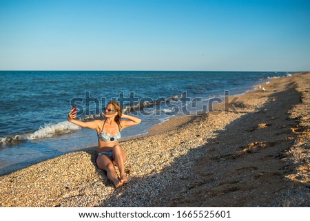 Beautiful young woman in a swimsuit makes selfie on a smartphone while relaxing on the beach by the sea on a sunny warm summer day. Social networking and leisure concept. Copyspace