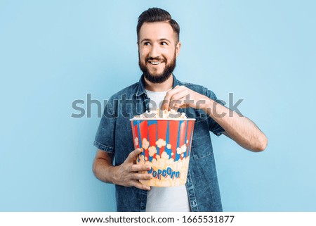 Happy bearded man holding a bucket of popcorn, spending an evening in a cozy home environment, young emotional guy watching a movie, on an isolated blue background