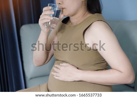 Close-up pregnant woman holding  glass of water sitting on sofa in the room. Nutrition and diet during pregnancy.