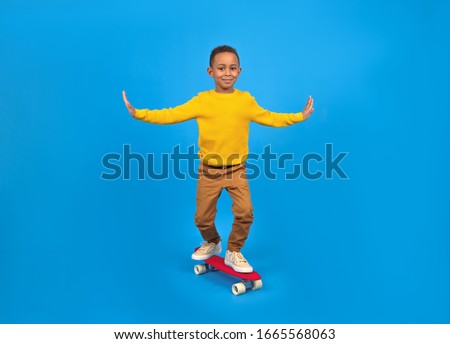 Happy boy is African American riding a red skateboard balancing his arms placed in sides for a balance-clad yellow sweater and brown pants against a blue studio background. free space for text
