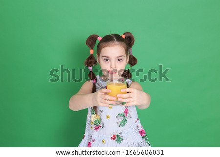Cool young beautiful girl of eight years old in excellent mood with a funny hairstyle in a blue blouse with flowers drinks orange juice from a transparent glass on a green background