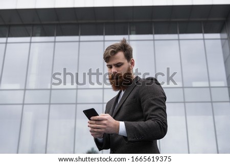 Young bearded Businessman holding mobile smartphone using app texting sms message wearing jacket outdoor. Successful entrepreneur dressed in formal wear communicating with a business partner online.
