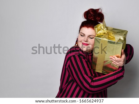 Happy ginger woman in black and purple striped dress, crown and earrings. Holding golden gift box tied with ribbon and bow, looking joyful, posing isolated on white. Close up, copy space