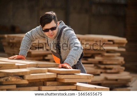 Portrait of young man carpenter at the wooden warehouse try to arrange or to use a wooden plank/board from a big wood timber stack of wooden planks.