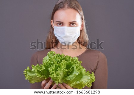 Portrait of a young woman in a mask and a knitted scarf, looking at a fresh green salad. The concept of protection against viruses during the flu epidemic, vitamins for health