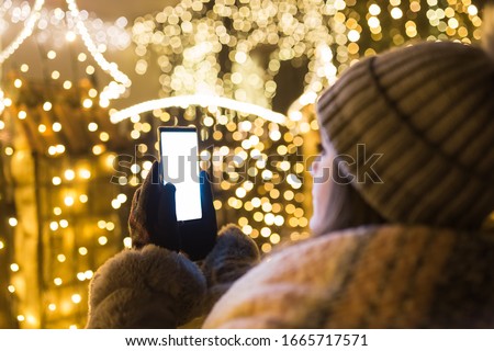 Woman using empty screen smart phone in front of house with new year christmas decoration lamps.