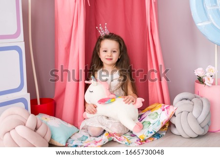 A little girl in a smart dress on the background of a beautiful children's room. Toy unicorn in his hands. Image of a little Princess in a crown