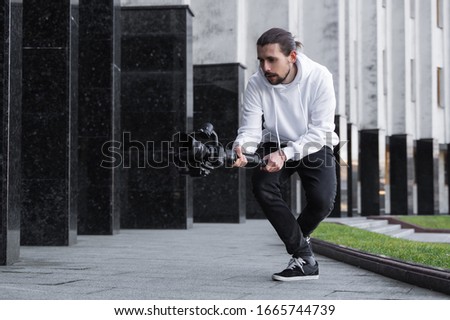Young Professional videographer holding professional camera on 3-axis gimbal stabilizer. Pro equipment helps to make high quality video without shaking. Cameraman wearing white hoodie making a videos. Royalty-Free Stock Photo #1665744739
