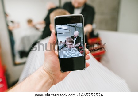 The camera on the buffer shoots a young man and a barber as he creates a stylish hairstyle. Man holds in his hand phone with camera makes photo of barber clipping client.