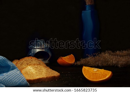 Still life in blue and yellow tones in a low key. Sliced bread, towel, orange, vase, lamp and reeds on a black background. Light brush technique. Focus in the background.