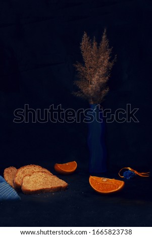 Still life in blue and yellow tones in a low key. Sliced bread, towel, orange, vase, lamp and reeds on a black background. Light brush technique. Focus in the background.