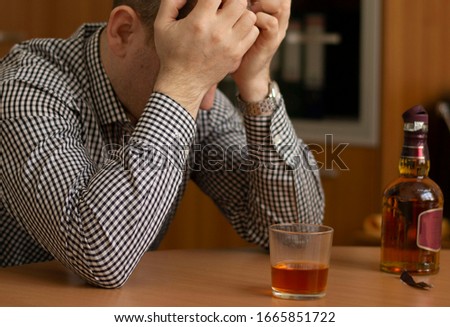 drunk man sitting at the table holding his head. on the table is a glass with alcohol and an open bottle