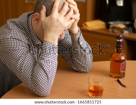drunk man sitting at the table holding his head. on the table is a glass with alcohol and an open bottle