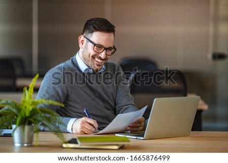 Portrait of young man sitting at his desk in the office. Handsome businessman in eyeglasses is using a computer while working in office. Young businessman using laptop at his office desk.