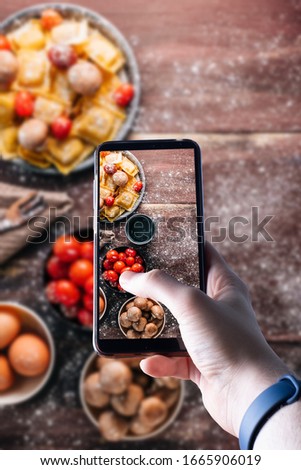 hands picking up a smartphone taking a picture of a fresh pasta on wooden table with ingredients. Space for text. Selective focus.