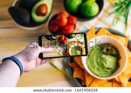 hands picking up a smartphone taking a picture of a bowl with guacamole. Space for text. Selective focus