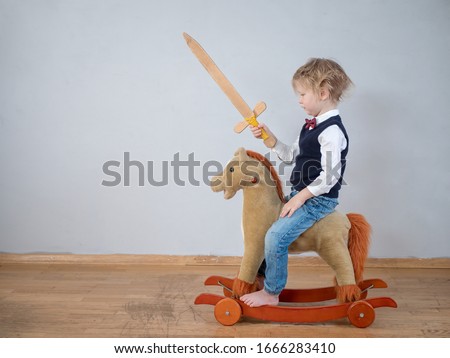Funny child in white shirt with bow-tie, on toy rocking horse with wooden sword. boy dreams of battles, victories and adventures. concept of education of spirit, education of morale, patriotism